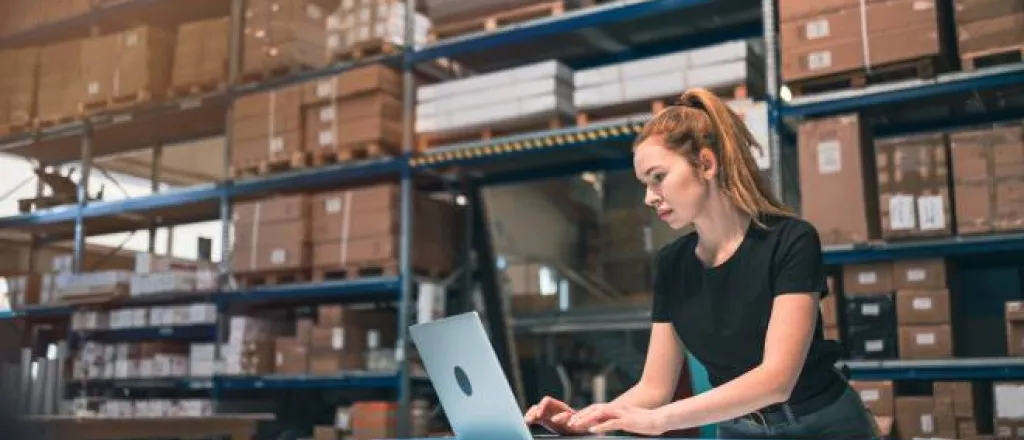 A woman with a ponytail wearing a black shirt standing in front of a table using a laptop in a warehouse.