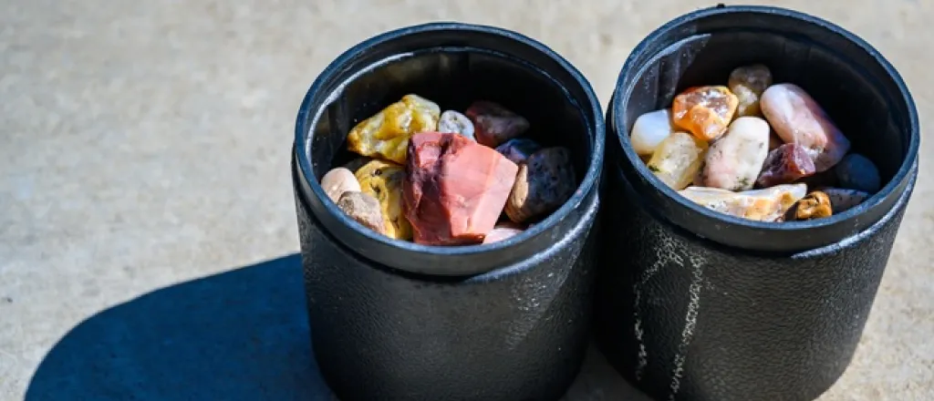 Two rubber rock tumbling barrels are placed on the ground and filled with different minerals in varying sizes.