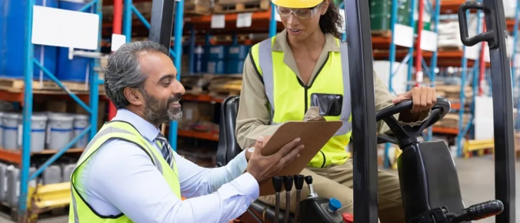 Two workers wearing safety vests. One is sitting in a forklift while the other is showing them information on a clipboard.