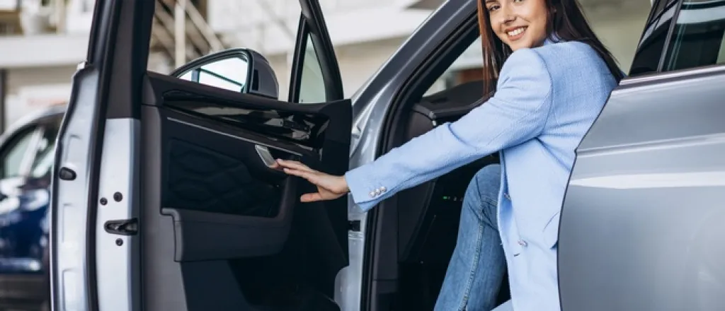 A woman in a blue suit jacket sits in the front seat of a silver car with the door open.