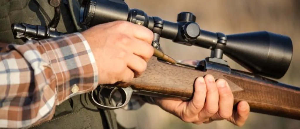  A close-up of a hunter, wearing a flannel shirt and vest, loading a round into a bolt-action carbine with a scope.