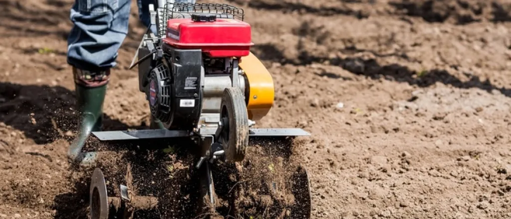 A person in farm attire walking a plot of land with a walking tiller. They're tilling the soil and wearing green boots.