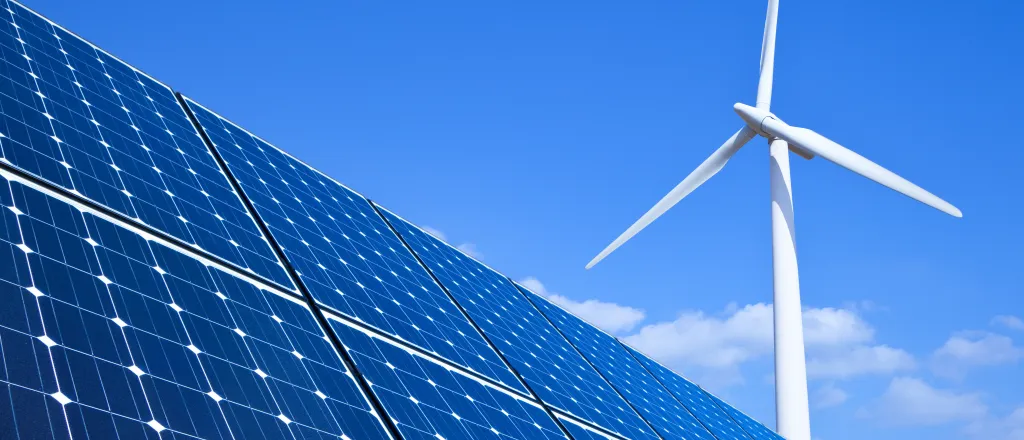 Solar panels in the foreground, with a wind turbine in the background, against a blue sky with a few clouds.