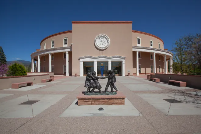 Entrance to the New Mexico State Capitol building. A statue of people is in a plaza area in front of the building.