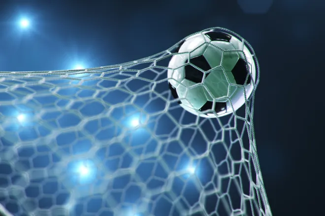 Soccer ball/football stretching the goal net with stadium lights in the background.
