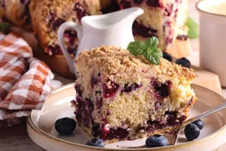 Plate with a slice of blueberry coffee cake with a few fresh blueberries. In the background are a small pitcher, additional slices of cake, and a cloth.
