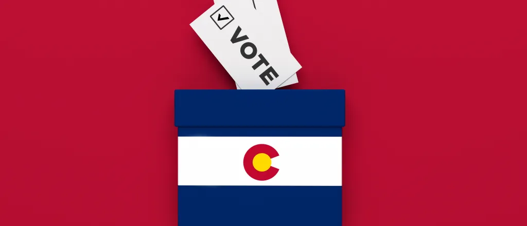Miniature ballot box with and image of the Colorado state flag on a red background with slips of paper representing ballots