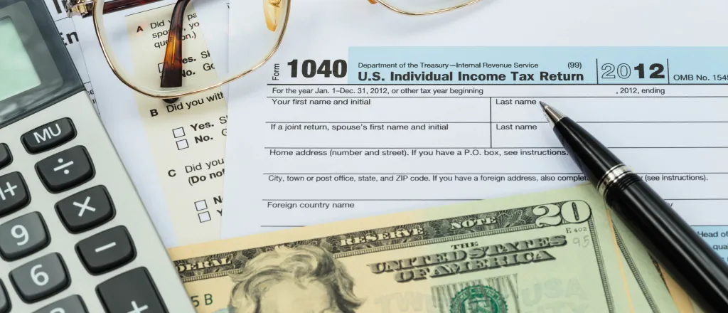 Closeup of United States currency, calculator, pen, and glasses on top of tax forms.