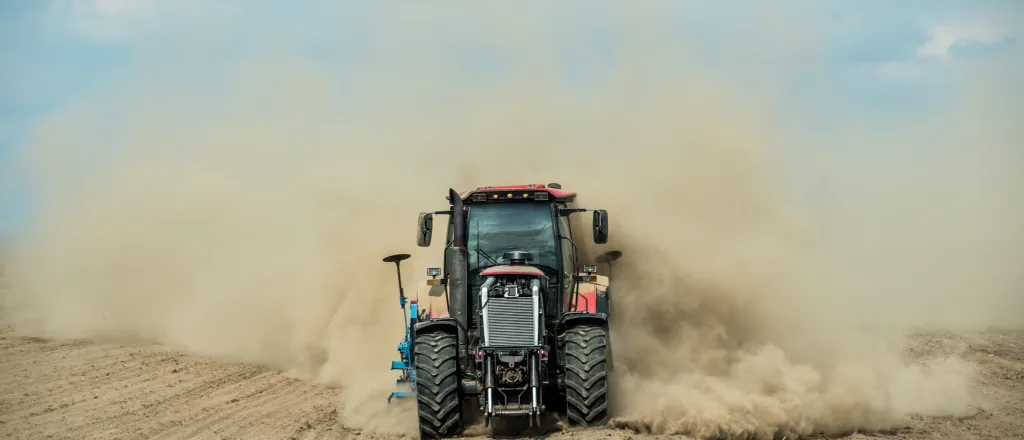Farm tractor driving through bare dirt creating a dust cloud.