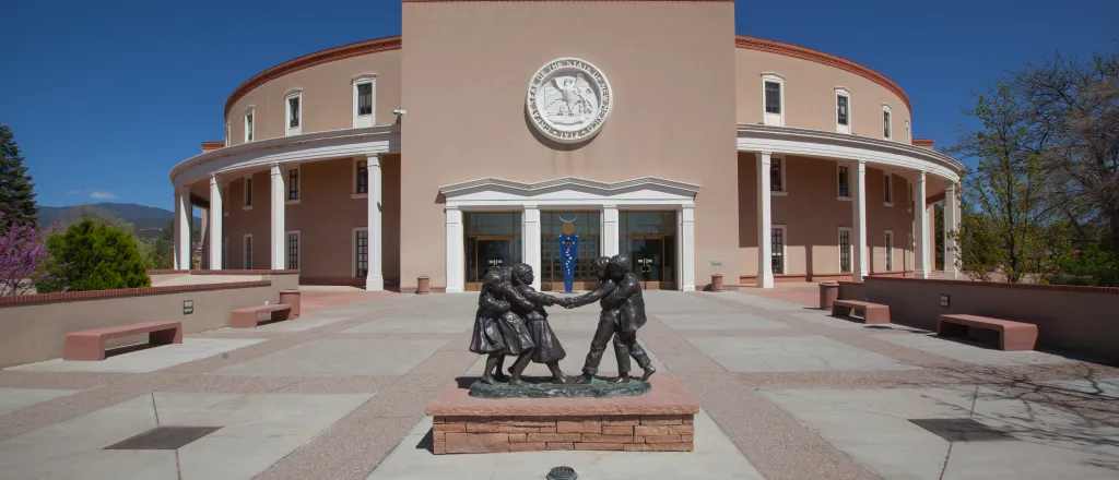 Entrance to the New Mexico State Capitol building. A statue of people is in a plaza area in front of the building.
