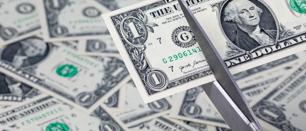 A pair of scissors is preparing to cut a United States one dollar note. In the background, additional dollar bills are strewn haphazardly.