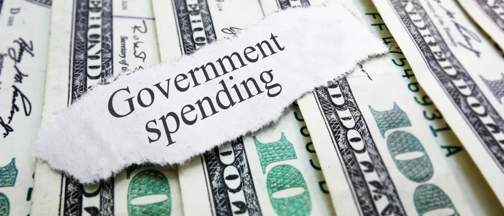 A scrap of torn paper with the words "Government spending" rests on top of a spread of United States 100 dollar currency.
