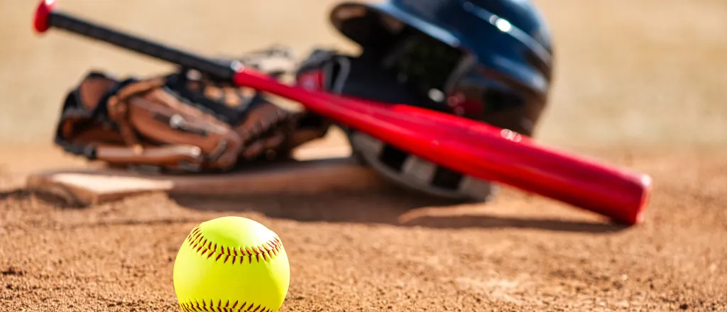 A softball sit in the foreground, with a helmet, bat, and glove sitting on a base slightly blurred in the background.
