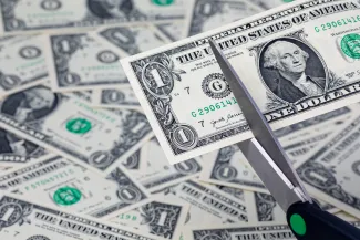 A pair of scissors is preparing to cut a United States one dollar note. In the background, additional dollar bills are strewn haphazardly.