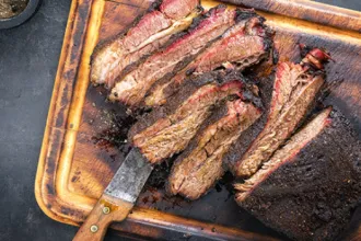 Overhead view of a tray of several slices of slow-cook barbecue. A knife sits under one slice.