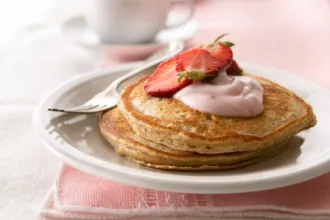 Plate on a placemat with two whole grain strawberry pancakes topped with a sliced fresh strawberry and yogurt. A fork rests on the plate. In the background a cup sits on a saucer.