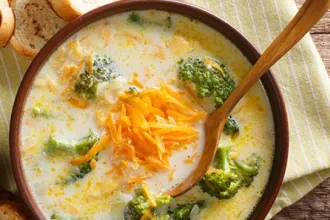 Overhead view of a bowl of broccoli potato soup. Shredded cheese rests on top of the soup. A wooden spoon is in the soup.